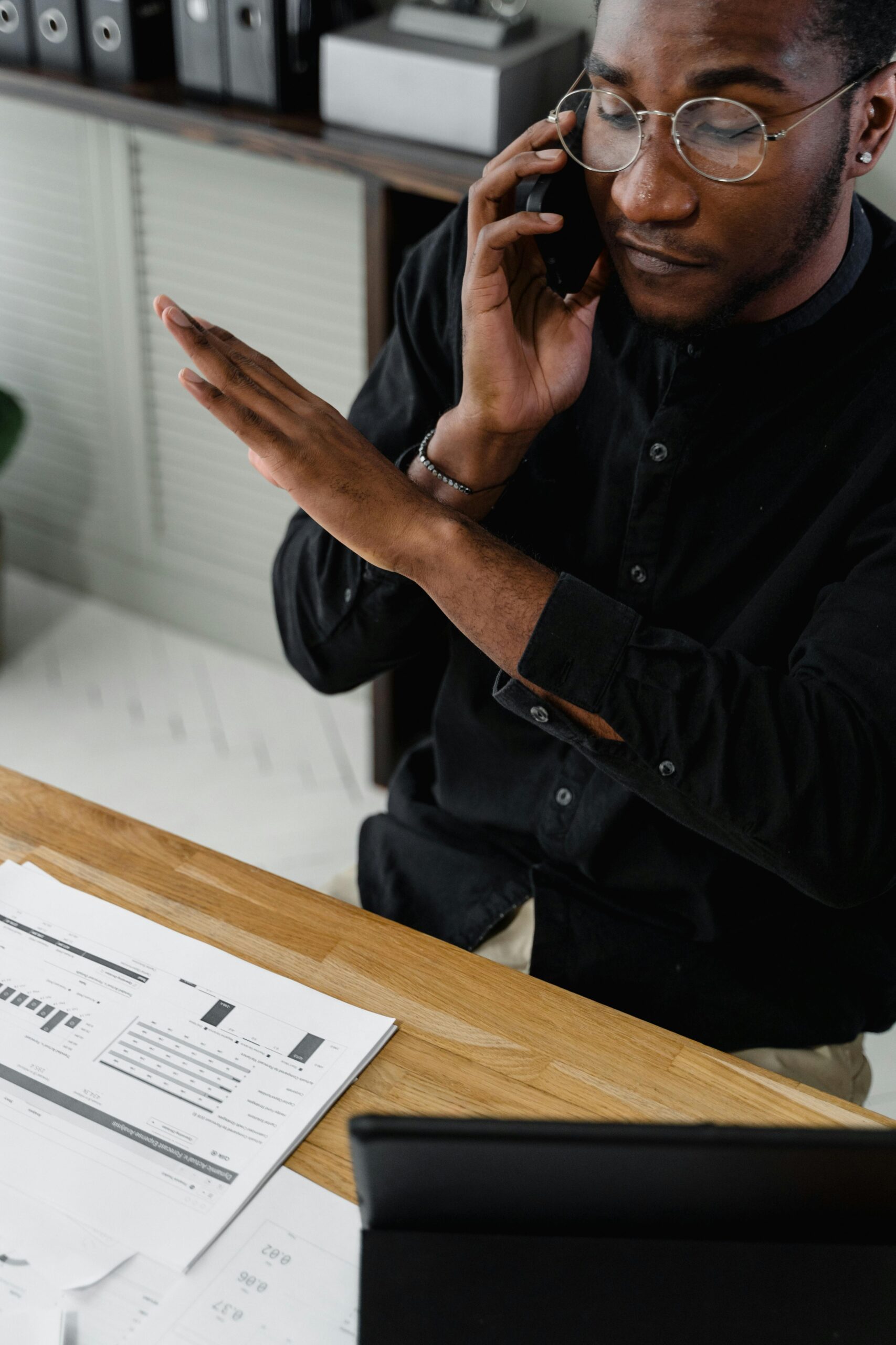 Black male accountant discussing financial reports on phone in office setting.